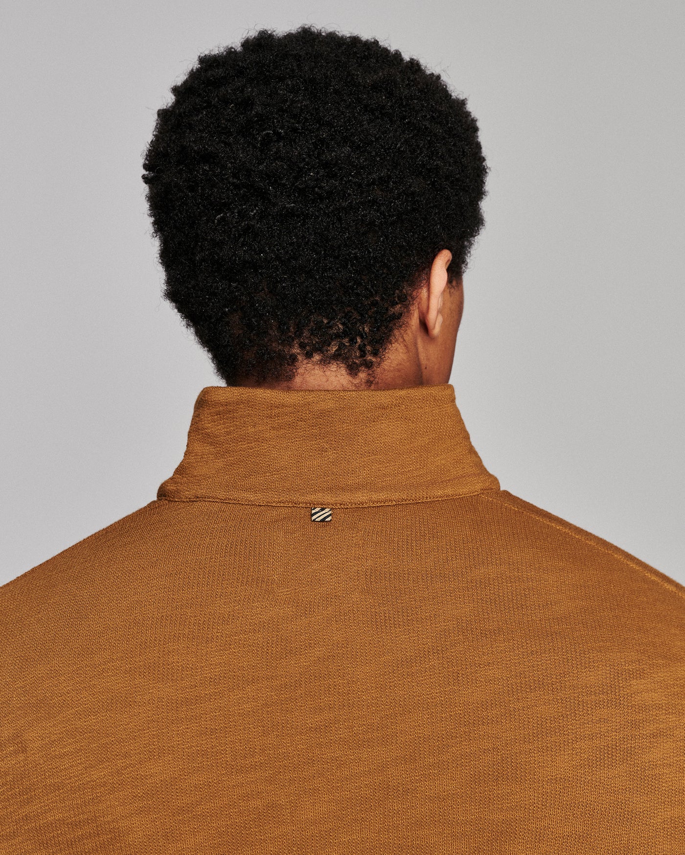 Back view of a person with curly black hair wearing a mustard brown ribbed knit mock neck sweater against a light gray background.