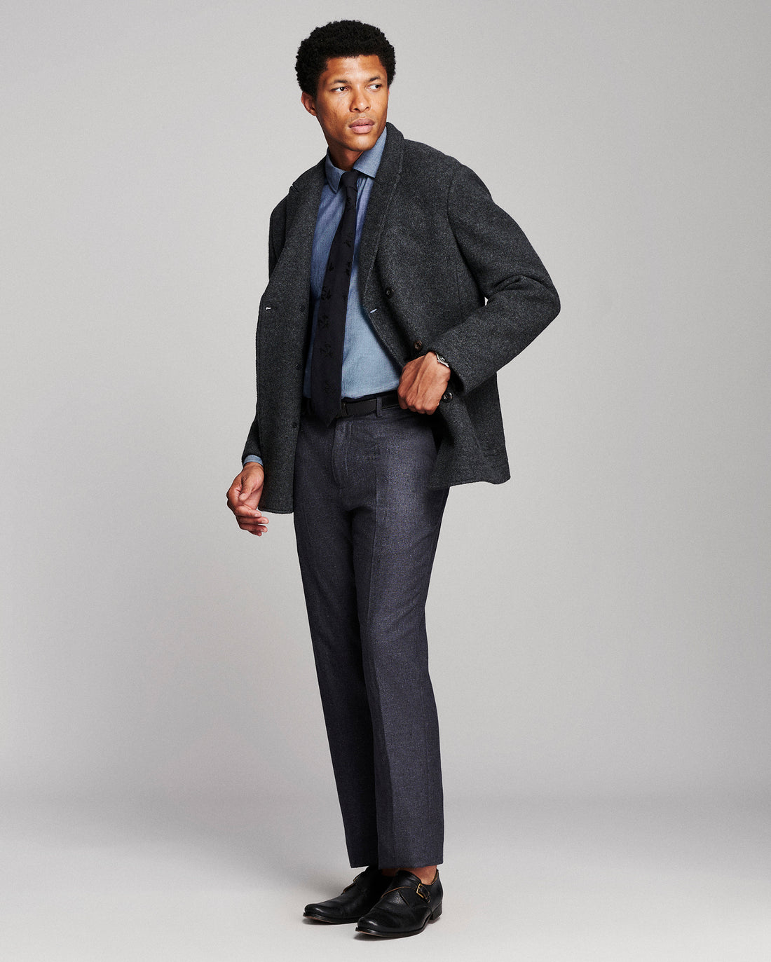 A man modeling a dark charcoal tweed blazer, dark gray trousers, a light blue dress shirt, a navy tie, and black dress shoes against a light gray studio background.