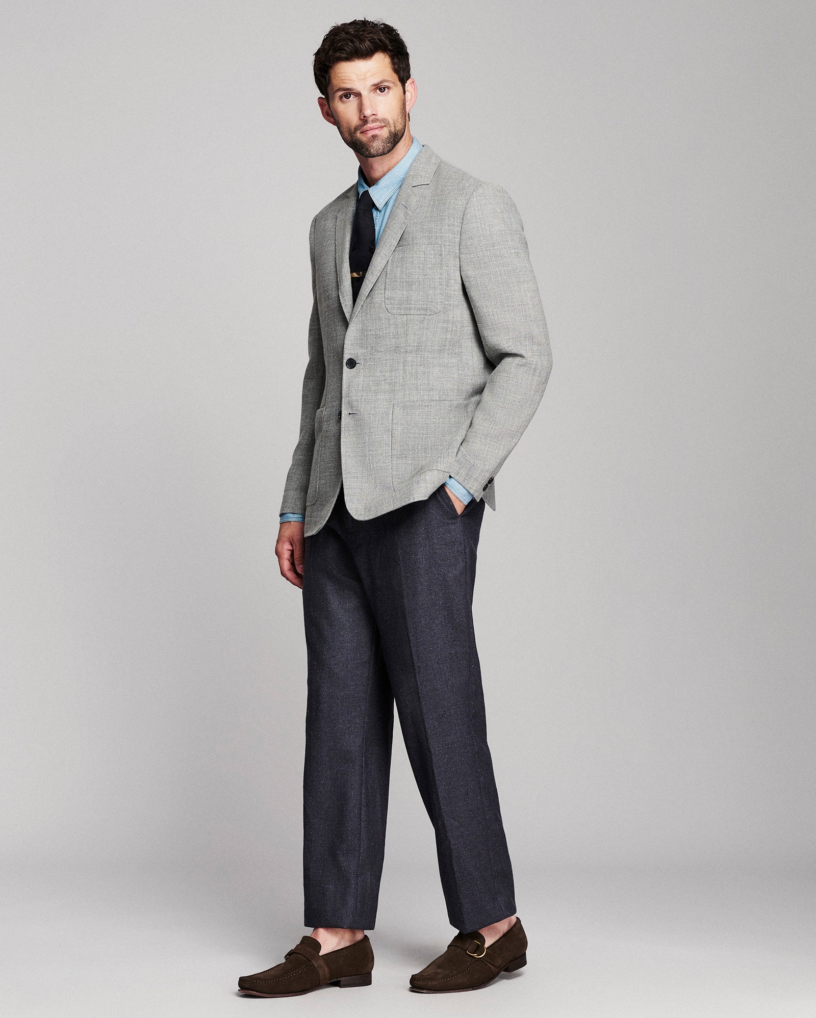 A man modeling a light gray two-piece suit with a subtle houndstooth pattern, featuring a light blue dress shirt, a dark tie, and dark brown loafers, set against a solid light gray studio background.