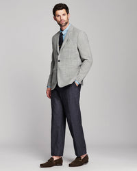 A man modeling a light gray two-piece suit with a subtle houndstooth pattern, featuring a light blue dress shirt, a dark tie, and dark brown loafers, set against a solid light gray studio background.