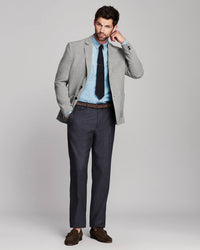 A man modeling a light gray blazer, light blue dress shirt, black tie, dark gray trousers, and brown loafers against a light gray studio background.