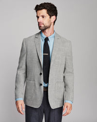 A man wearing a light gray textured blazer over a light blue dress shirt and a black tie, paired with dark trousers, photographed against a solid gray studio background.