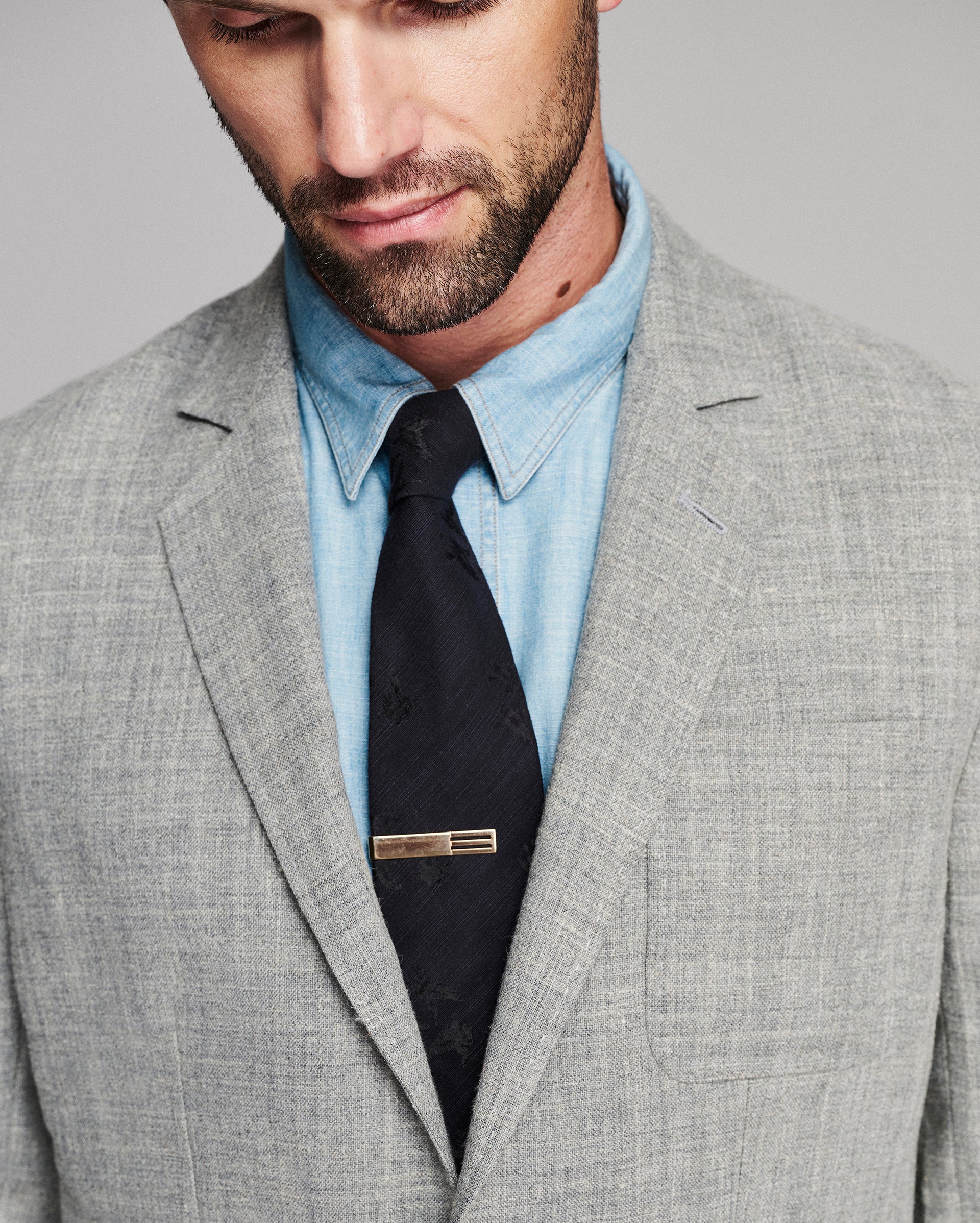 A close-up of a man wearing a light gray textured suit jacket, a light blue dress shirt, a black tie, and a gold-toned tie clip against a neutral gray background.