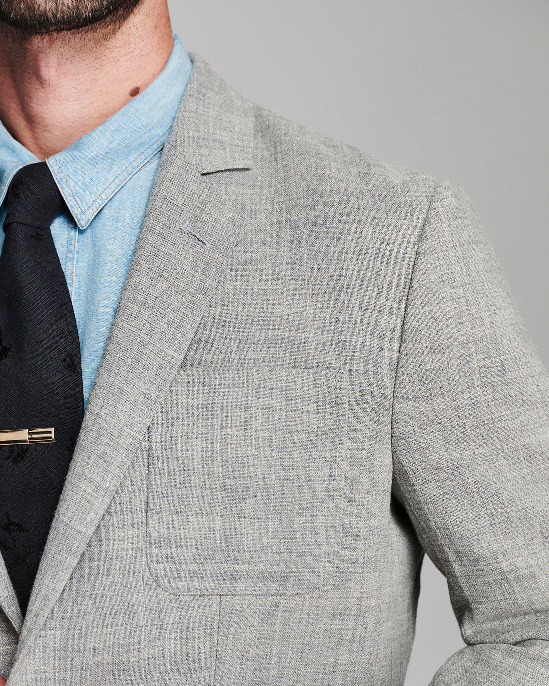 Close-up of a man wearing a light gray textured blazer over a light blue dress shirt and a black tie with a gold tie bar, against a solid gray background.