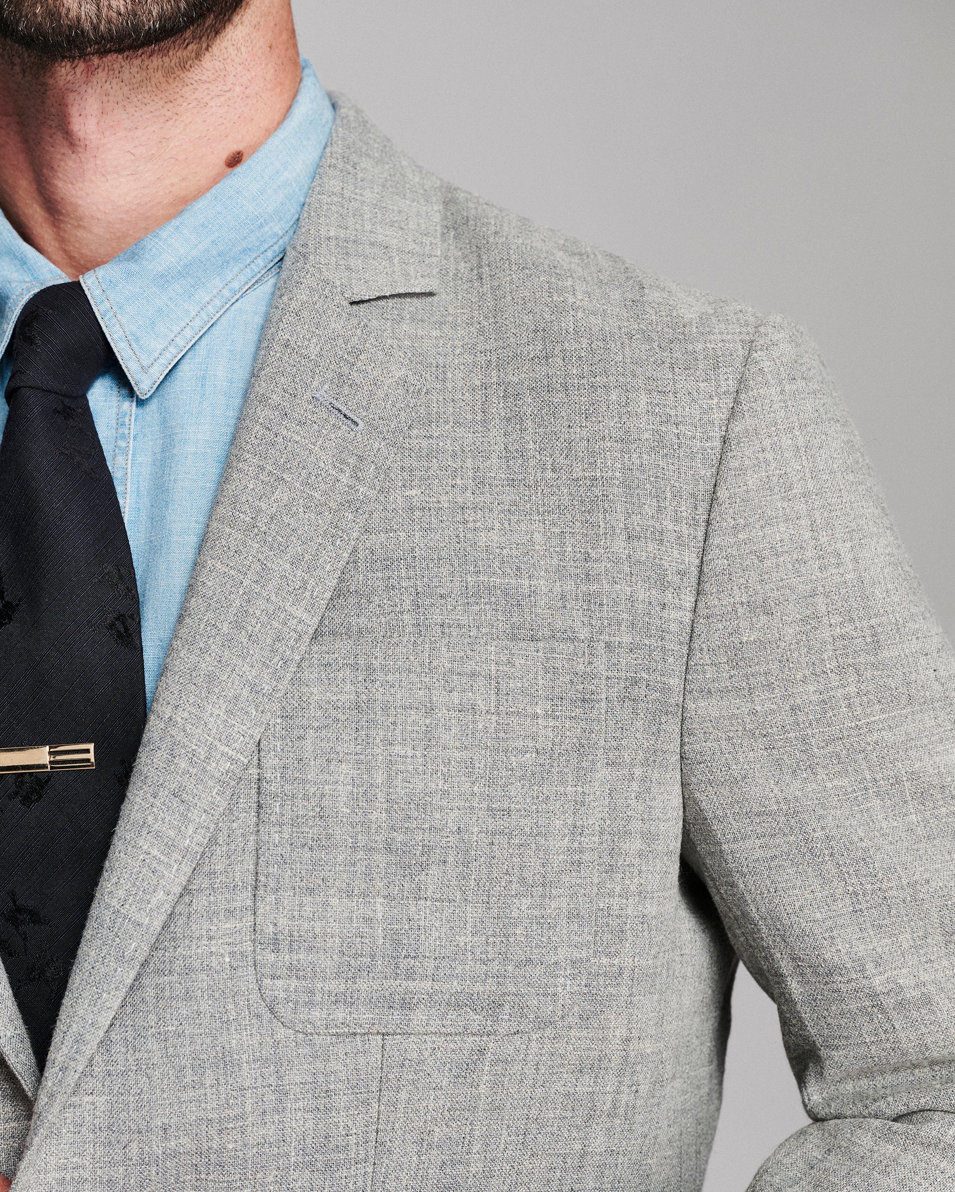 Close-up of a man wearing a light gray textured blazer over a light blue dress shirt and a black tie with a gold tie bar, against a solid gray background.