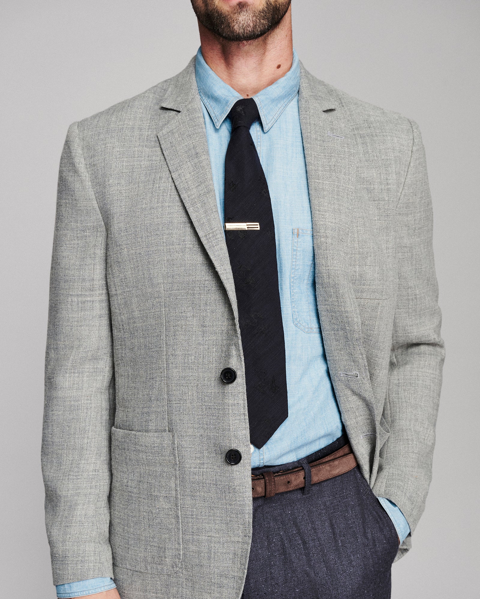 A man wearing a light gray textured blazer over a light blue dress shirt and black tie, paired with dark gray trousers and a brown belt, photographed against a neutral gray studio background.