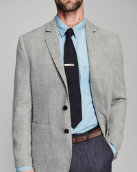 A man wearing a light gray textured blazer over a light blue dress shirt and black tie, paired with dark gray trousers and a brown belt, photographed against a neutral gray studio background.