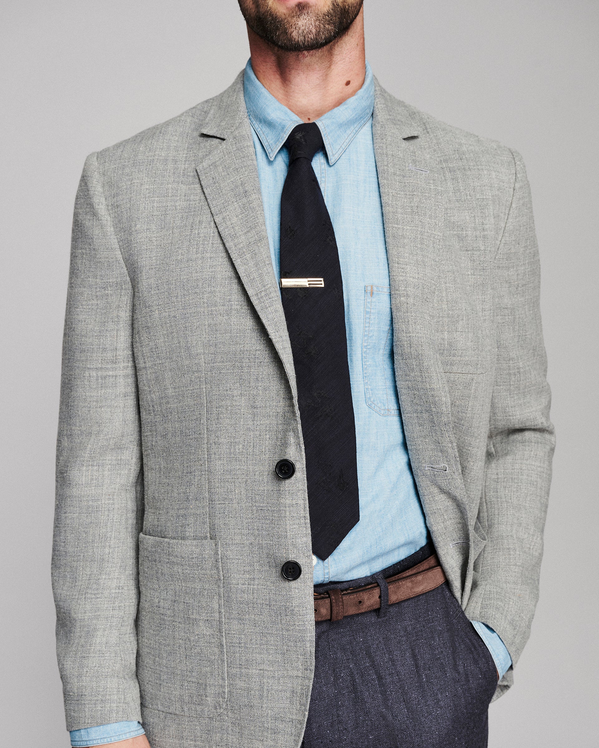 A man wearing a light gray textured blazer over a light blue dress shirt and black tie, paired with dark gray trousers and a brown belt, photographed against a neutral gray studio background.
