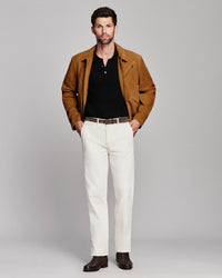 A man modeling a brown suede jacket over a black henley shirt, paired with off-white trousers and dark brown leather shoes, against a solid light gray studio background.