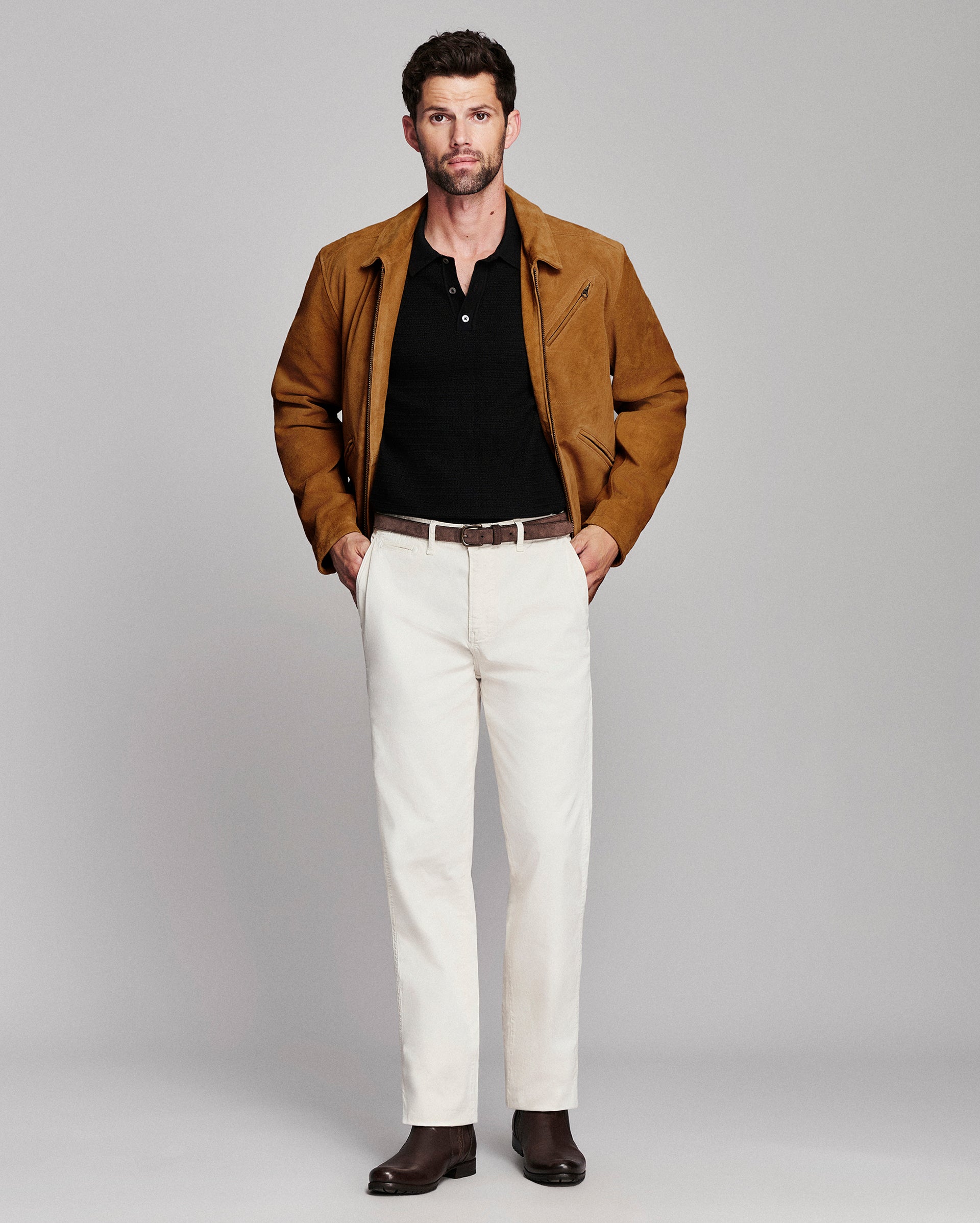 A man modeling a brown suede jacket over a black henley shirt, paired with off-white trousers and dark brown leather shoes, against a solid light gray studio background.