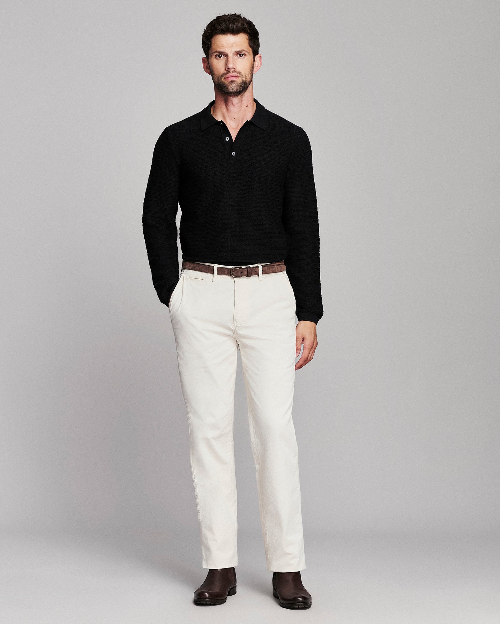 A man modeling a black long-sleeve polo shirt with white trousers, a brown belt, and dark brown shoes, standing against a light gray studio background.