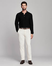 A man modeling a black long-sleeve polo shirt with white trousers, a brown belt, and dark brown shoes, standing against a light gray studio background.