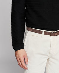 Close-up of a person wearing a black ribbed knit sweater, off-white trousers, and a brown belt against a light gray studio background.
