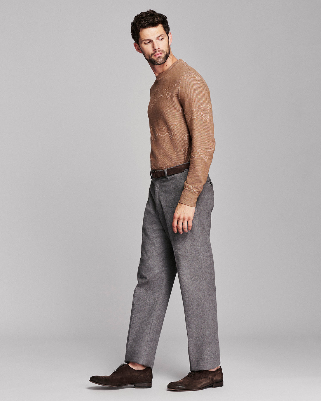 A man modeling a brown knit crewneck sweater with a subtle tonal pattern, paired with gray trousers and dark brown suede loafers, shot against a solid gray studio background.