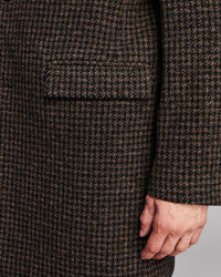 Close-up of a dark brown and black houndstooth patterned coat with a hand resting near the hem, showing a pocket and fabric texture.