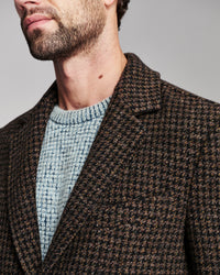 Close-up of a man wearing a brown and black houndstooth blazer over a light blue knit sweater, against a neutral gray background.