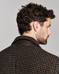 A man shown from the back and side profile wearing a dark brown and black houndstooth patterned jacket against a light gray studio background.