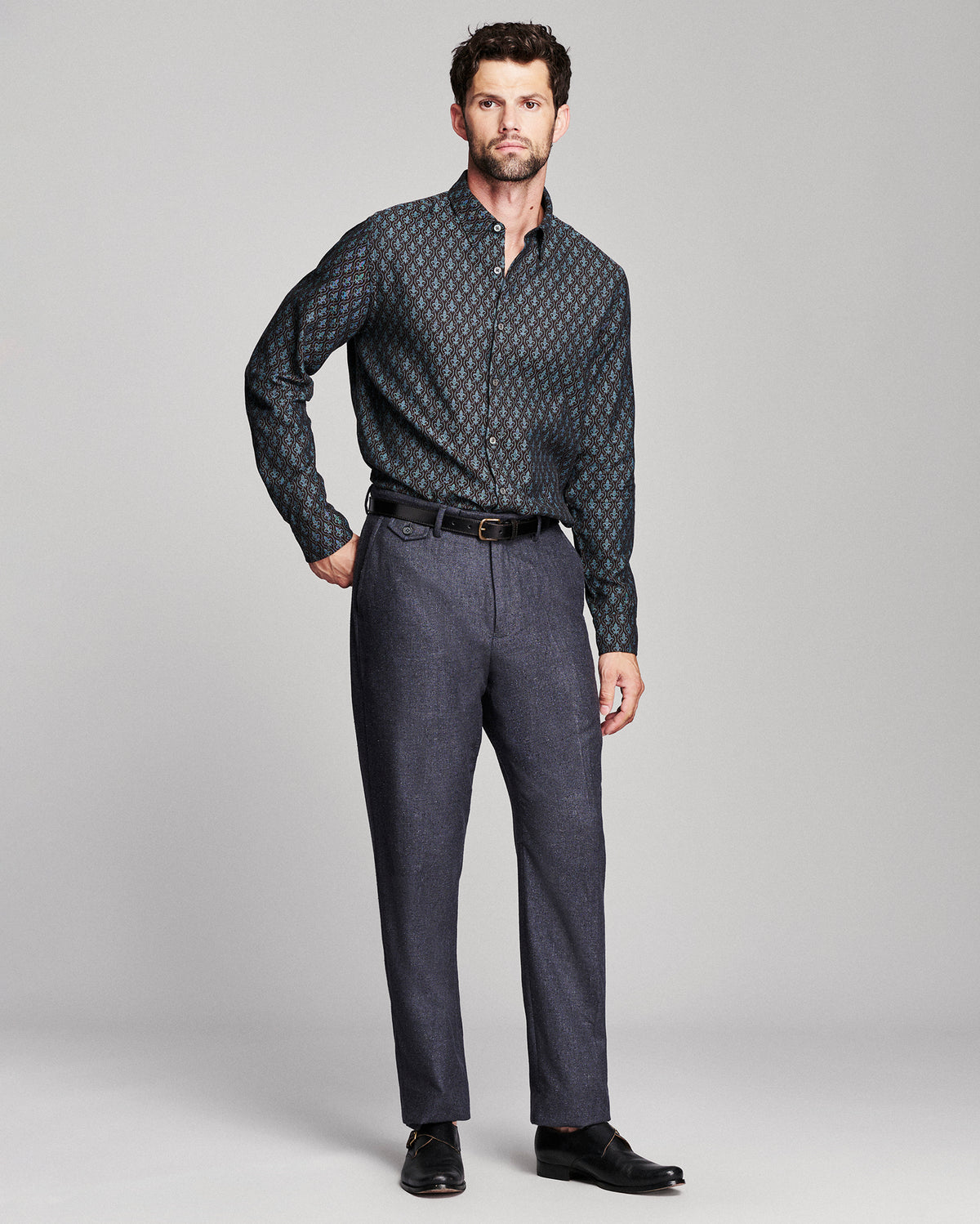 A man modeling a long-sleeve button-down shirt with a dark blue and black geometric pattern, paired with charcoal gray trousers, a black belt, and black dress shoes, against a light gray studio background.