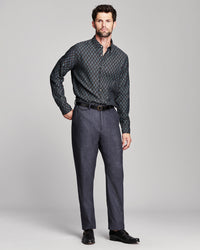 A man modeling a long-sleeve button-down shirt with a dark blue and black geometric pattern, paired with charcoal gray trousers, a black belt, and black dress shoes, against a light gray studio background.