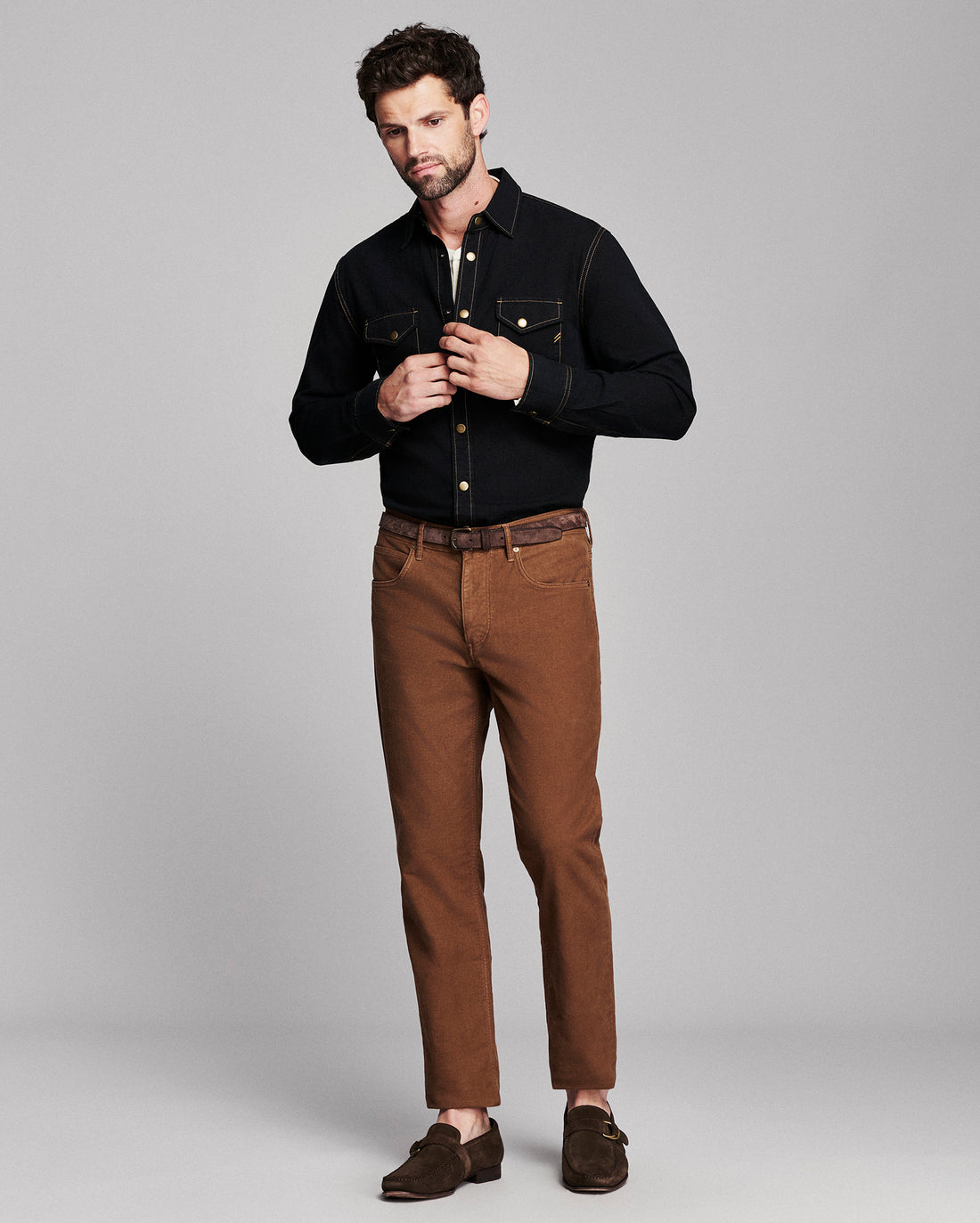 A man modeling a black long-sleeve button-up shirt and brown trousers against a solid gray studio background.