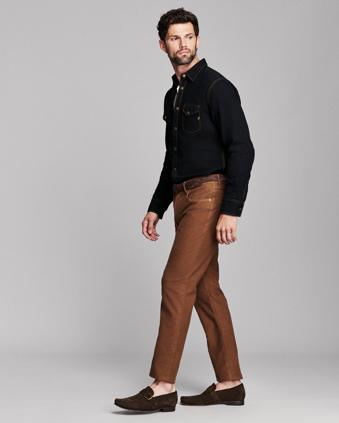 A man modeling a black long-sleeve button-up shirt and brown slim-fit jeans with brown leather loafers, standing against a light gray studio background.