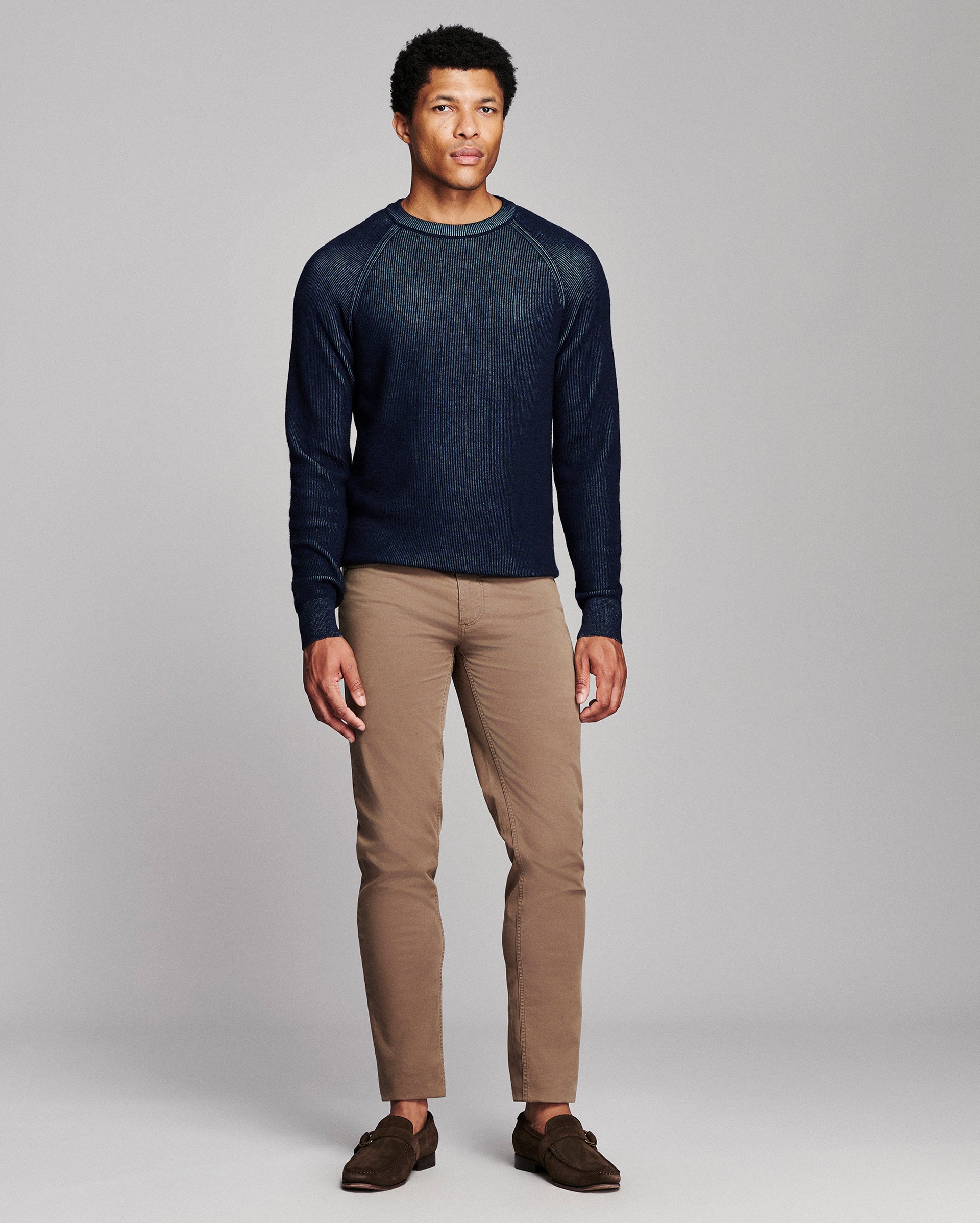 A man modeling a dark navy blue long-sleeve knit sweater with heathered texture, paired with tan pants and brown loafers, against a solid light gray studio background.