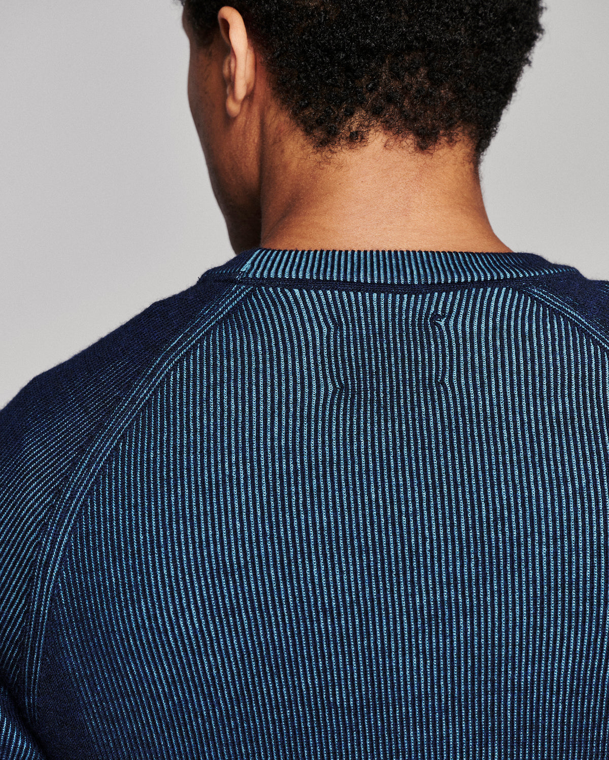Close-up back view of a person wearing a dark blue ribbed knit crewneck sweater against a light gray studio background.