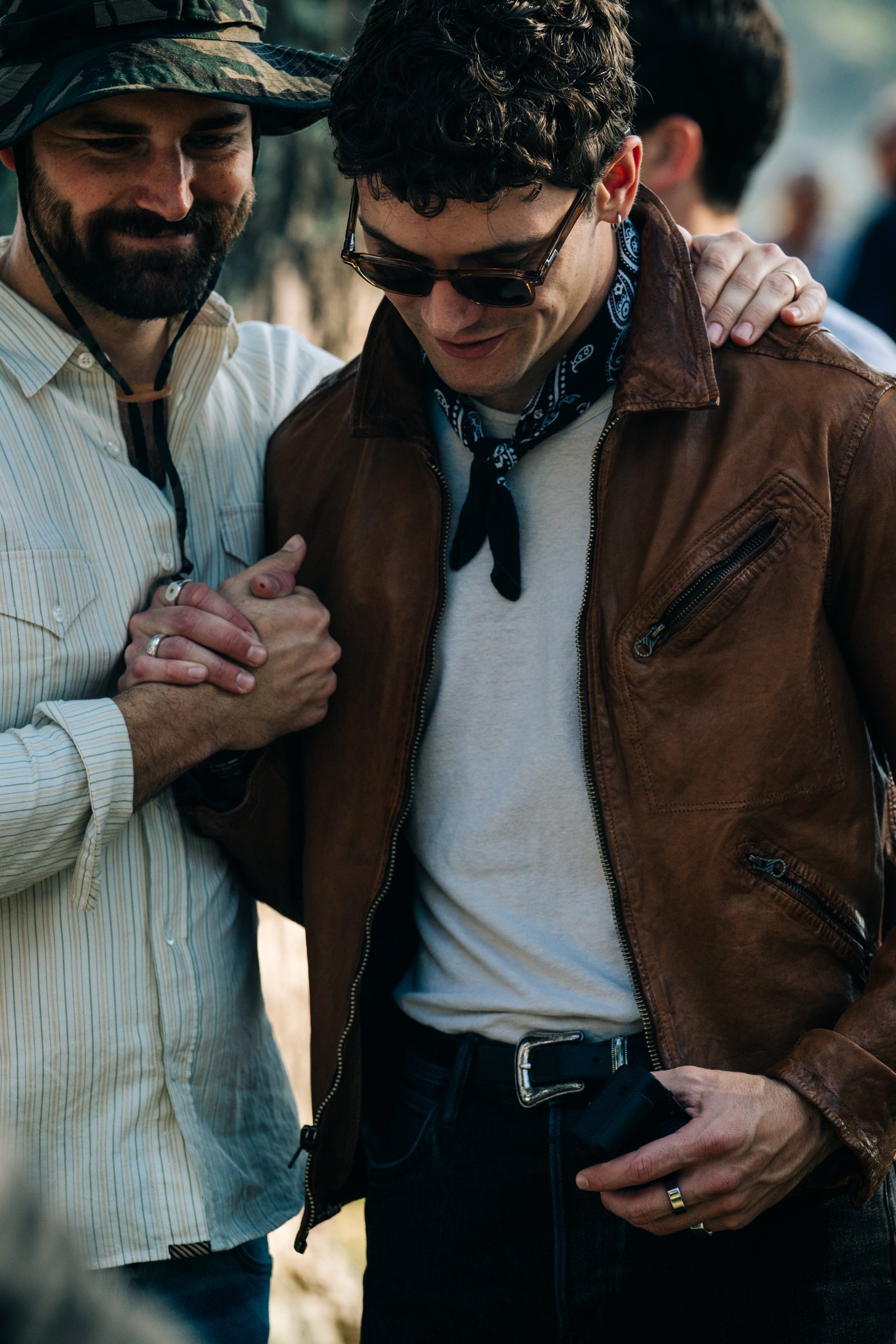 Two men standing close together outdoors. The man on the right wears a brown leather jacket, white shirt, black pants, sunglasses, and a black bandana.