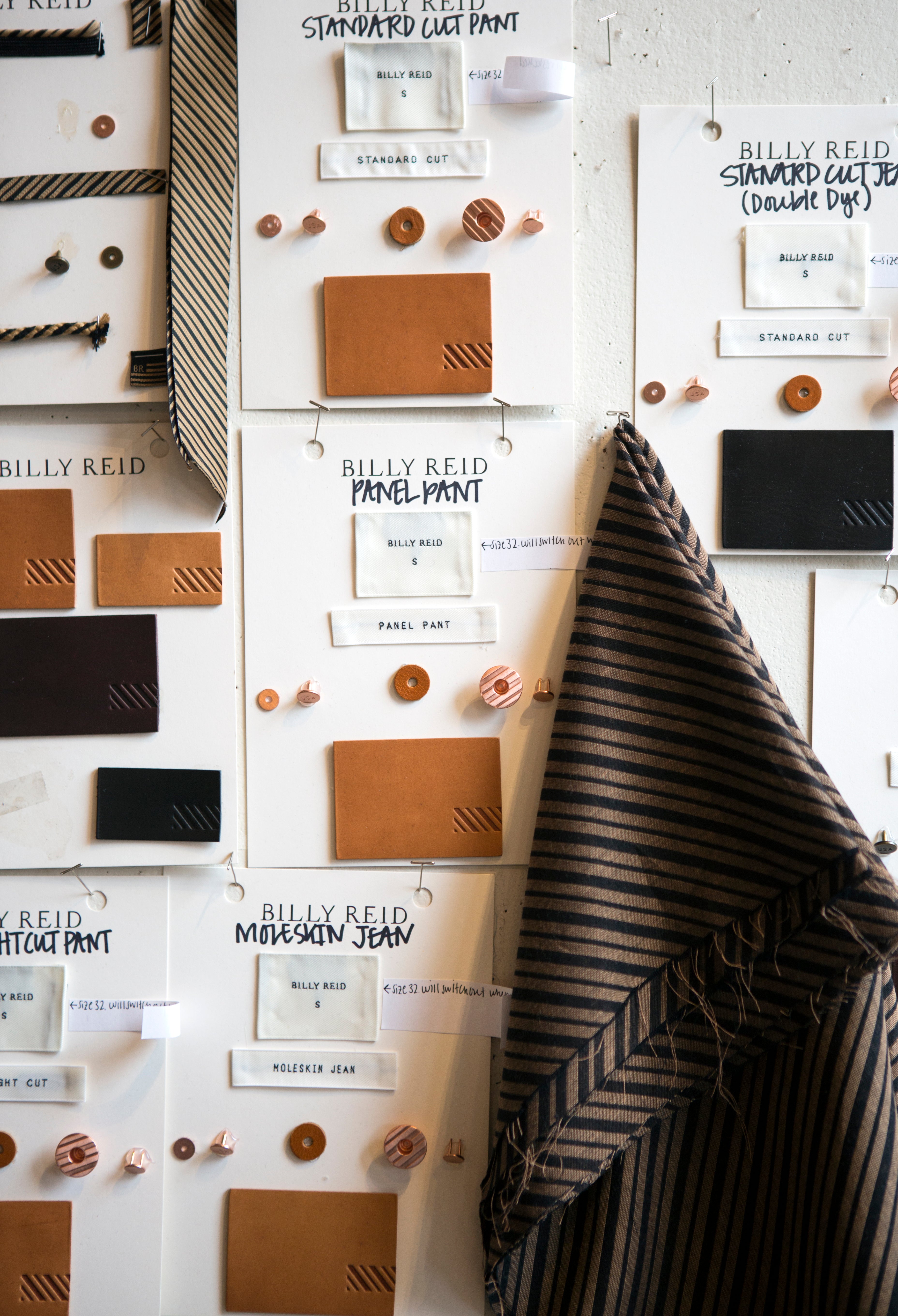 A styled flat-lay display of Billy Reid clothing materials and color swatches pinned to a white wall, featuring various shades of brown, beige, and black fabrics, leather panels, and fabric swatches labeled with different shirt and pant styles.