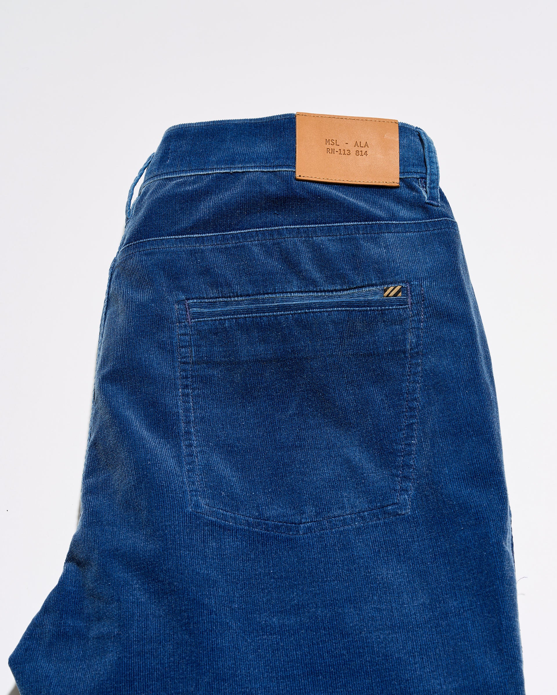Close-up of the back waistband and pocket area of blue corduroy jeans, showing a tan leather brand patch and a small yellow embroidered logo on the pocket.