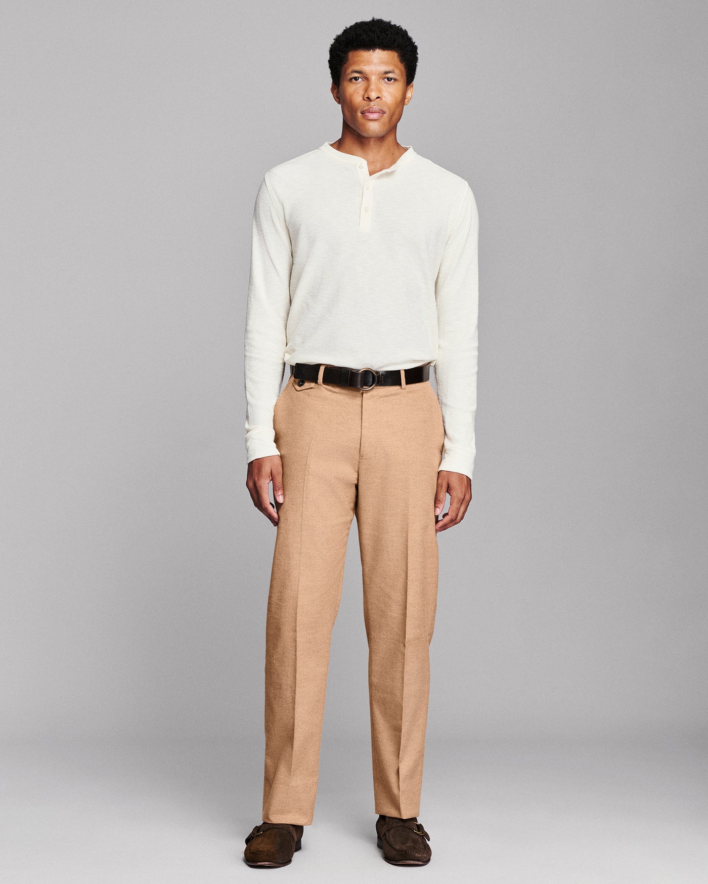 A man modeling a cream long-sleeve henley shirt and tan trousers with a black belt, standing against a solid gray studio background.