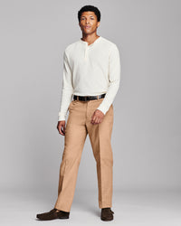 A man modeling a cream long-sleeve henley shirt, tan dress pants, a black belt, and dark brown loafers against a solid gray studio background.