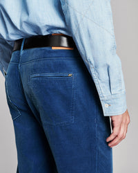 Close-up back view of a person wearing dark blue jeans, a light blue shirt, and a black belt against a plain light gray background.
