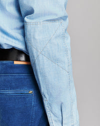 Close-up of a person wearing a light blue chambray long-sleeve shirt tucked into dark blue jeans with a black belt, showing the shirt's pocket and fabric texture.