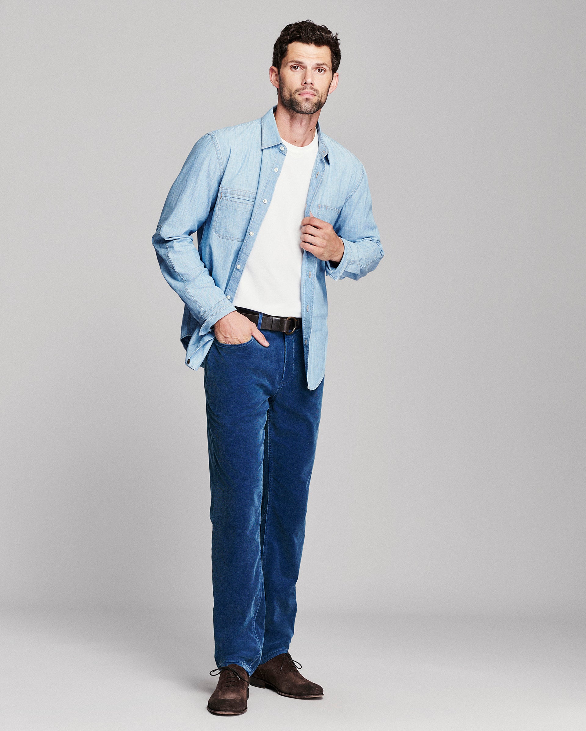 A man modeling a light blue denim shirt jacket over a white t-shirt, paired with dark blue corduroy trousers and brown leather shoes, against a solid light gray studio background.