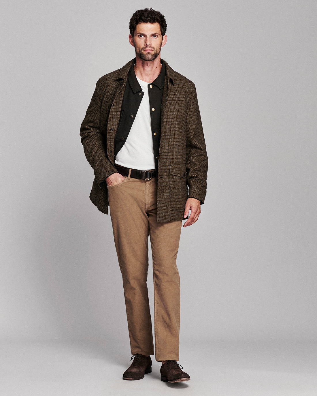 A man modeling a brown textured overshirt over a black button-up shirt and white tee, paired with tan trousers and dark brown leather shoes, against a solid gray studio background.