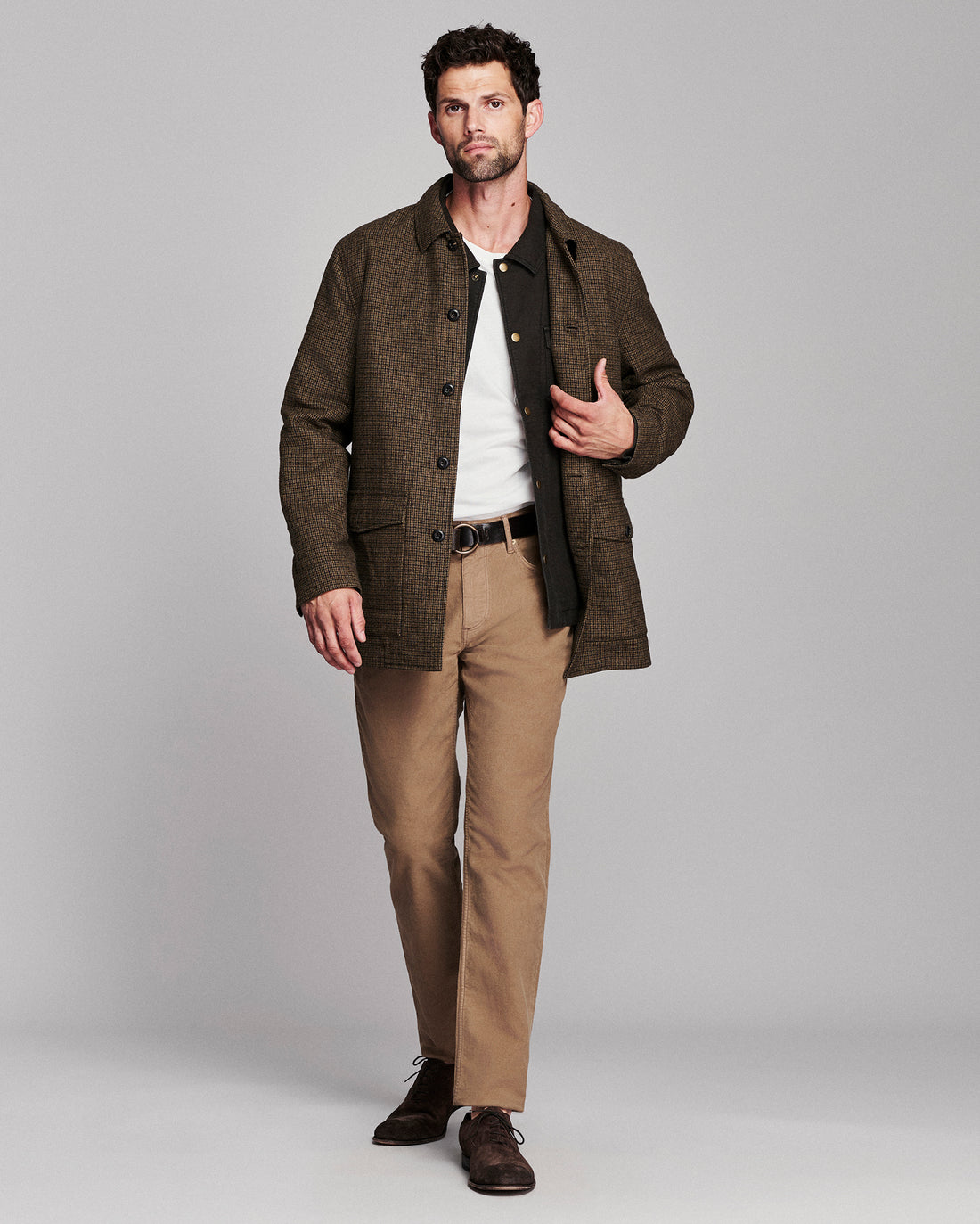 A man modeling a brown textured jacket over a white shirt and black vest, paired with tan trousers and dark brown shoes, against a solid gray studio background.