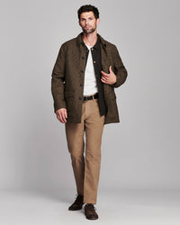 A man modeling a brown textured jacket over a white shirt and black vest, paired with tan trousers and dark brown shoes, against a solid gray studio background.