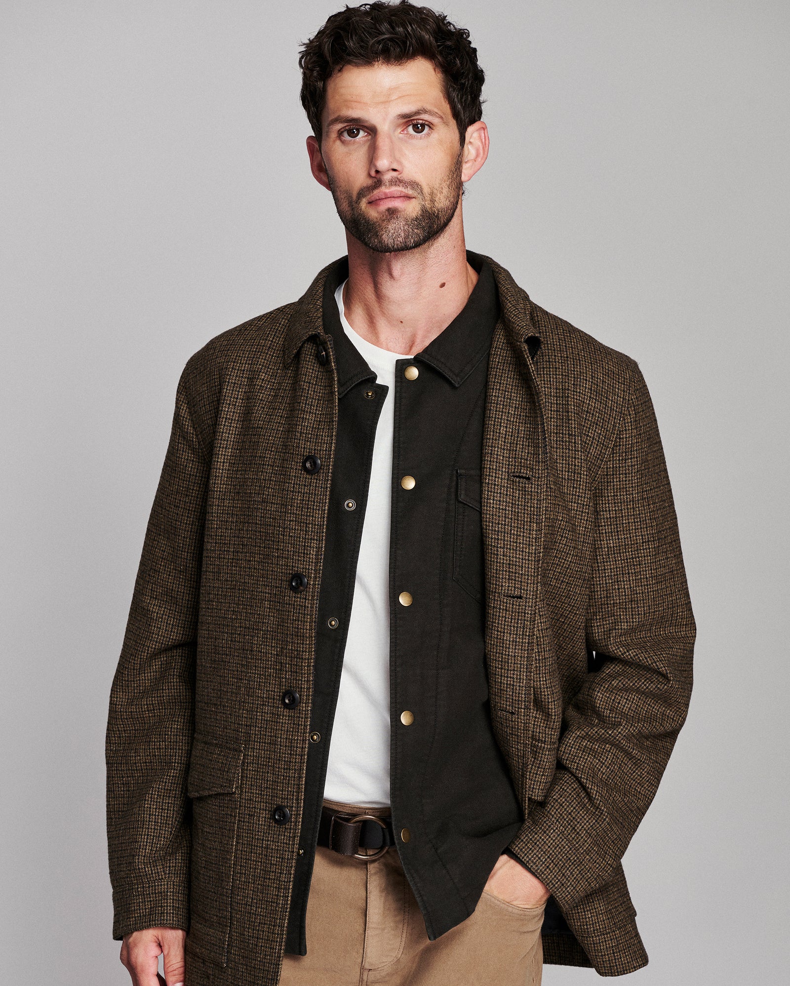 A man wearing a brown and black checkered overshirt over a black button-up shirt, white t-shirt, khaki pants, and a brown belt, photographed against a solid gray studio background.