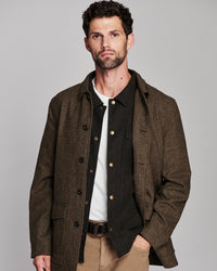 A man wearing a brown and black checkered overshirt over a black button-up shirt, white t-shirt, khaki pants, and a brown belt, photographed against a solid gray studio background.