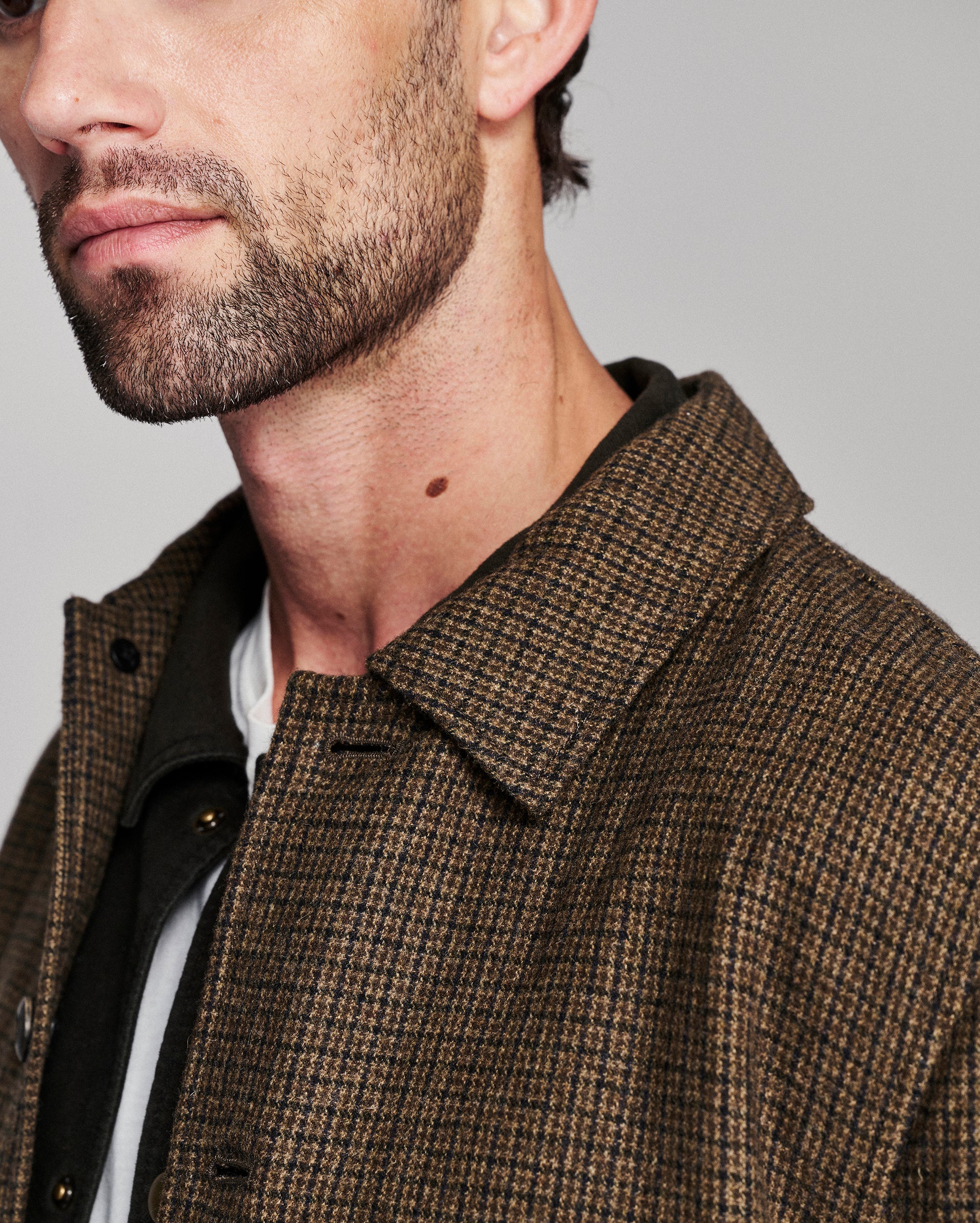 Close-up of a man wearing a brown and black houndstooth patterned overshirt over a black jacket and white shirt, against a neutral gray background.