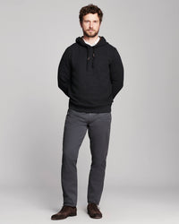 A man modeling a black textured knit hoodie with gray trousers and brown shoes, standing against a light gray studio background.