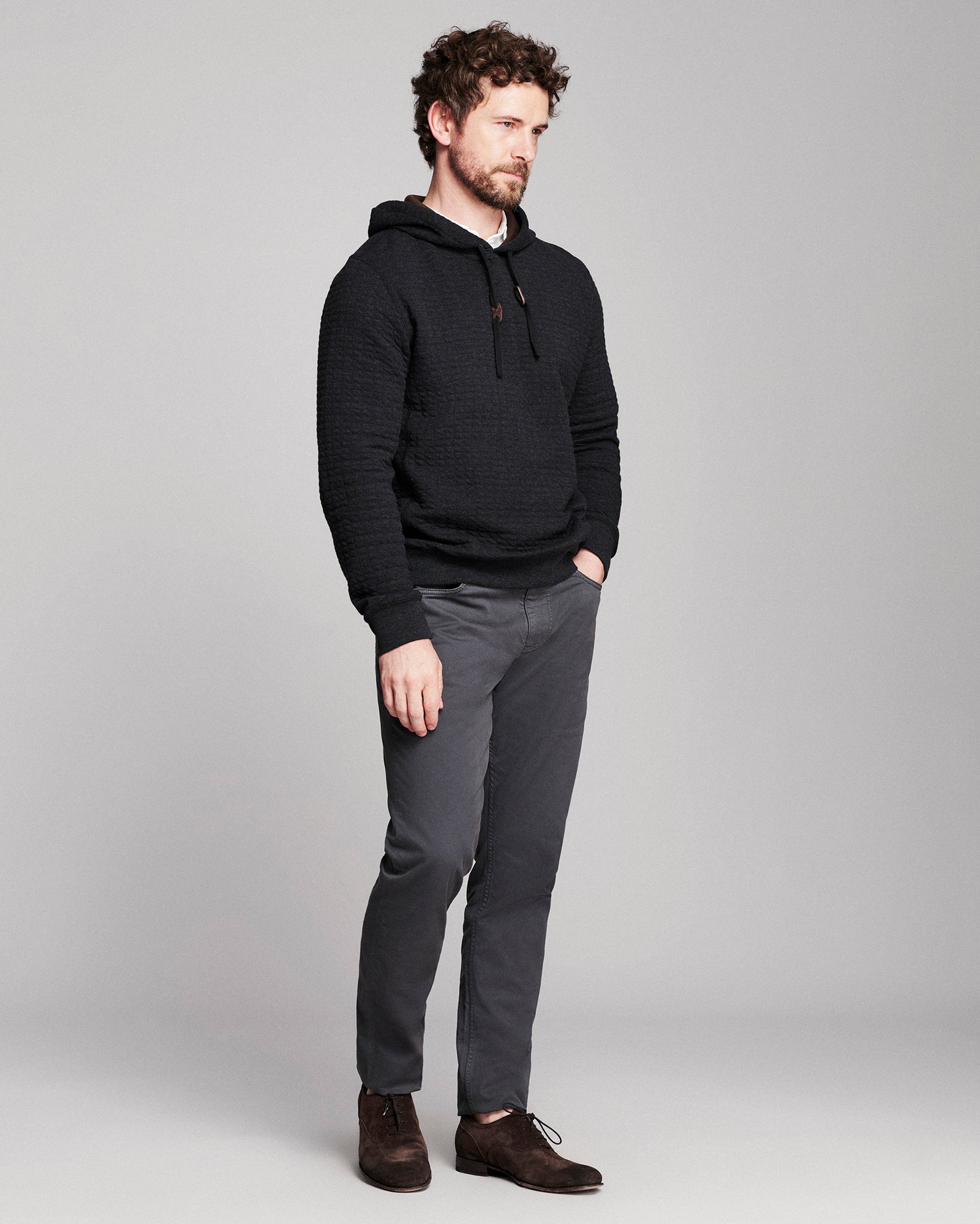 A man wearing a black textured pullover hoodie, dark gray slim-fit trousers, and brown leather shoes, standing against a light gray studio background.
