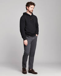 A man wearing a black textured pullover hoodie, dark gray slim-fit trousers, and brown leather shoes, standing against a light gray studio background.
