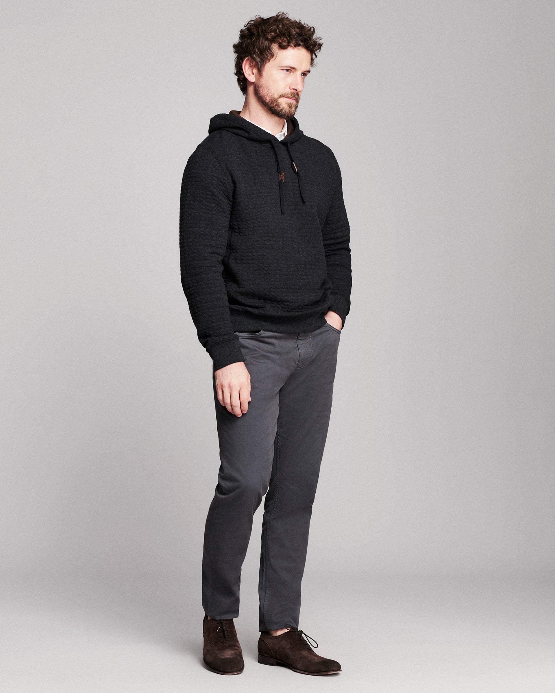 A man wearing a black textured pullover hoodie, dark gray slim-fit trousers, and brown leather shoes, standing against a light gray studio background.