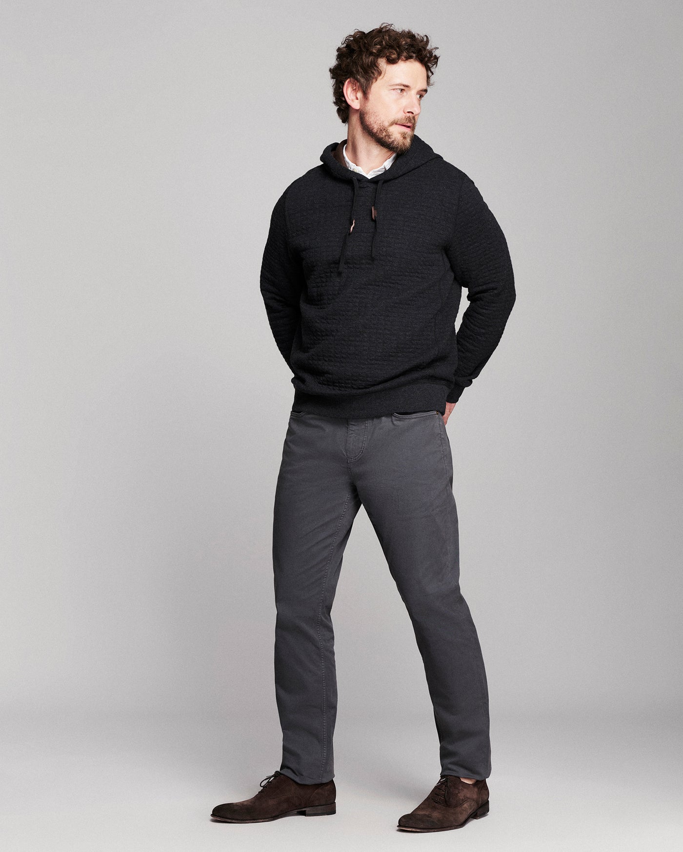 A man modeling a black textured knit hoodie and dark gray trousers against a light gray studio background.