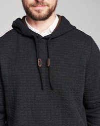 A close-up of a man wearing a dark charcoal gray hooded sweater over a white collared shirt, against a plain light gray background.