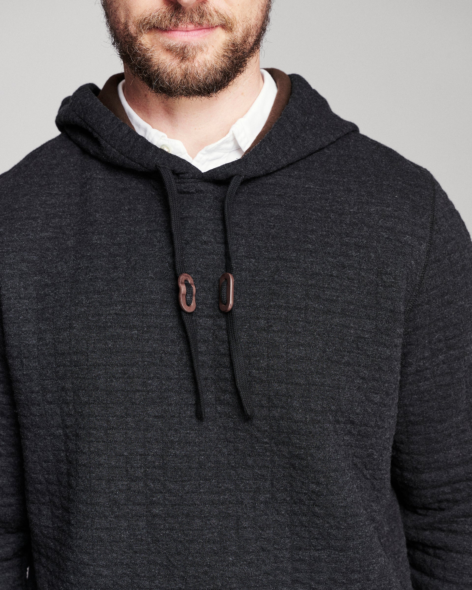 A close-up of a man wearing a dark charcoal gray hooded sweater over a white collared shirt, against a plain light gray background.