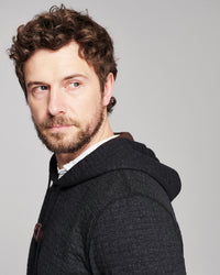 A man with curly brown hair and a beard, wearing a dark gray heathered hoodie over a white shirt, photographed against a solid light gray background.