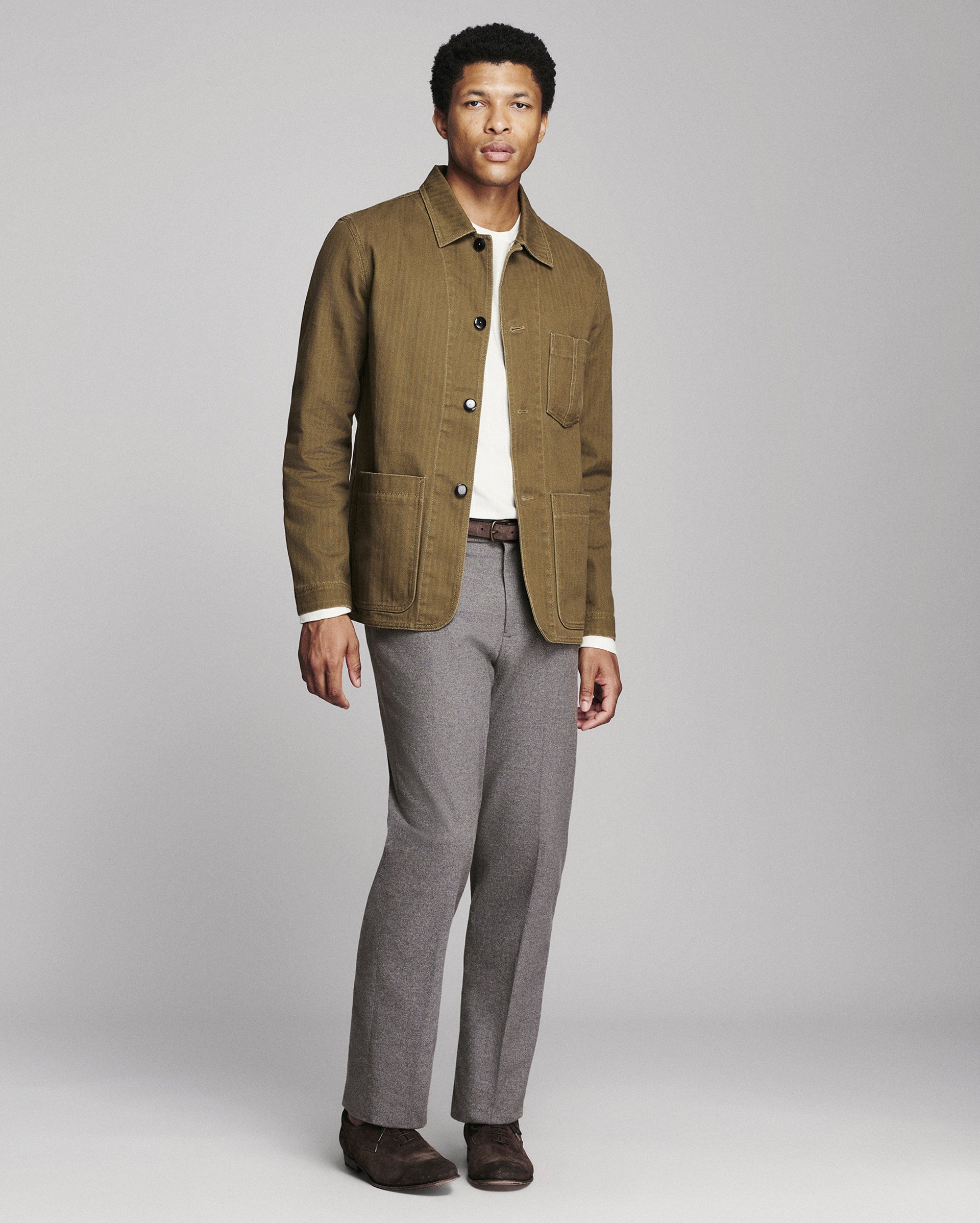 A man modeling a brown corduroy utility jacket over a white shirt, paired with gray trousers and brown shoes, against a light gray studio background.